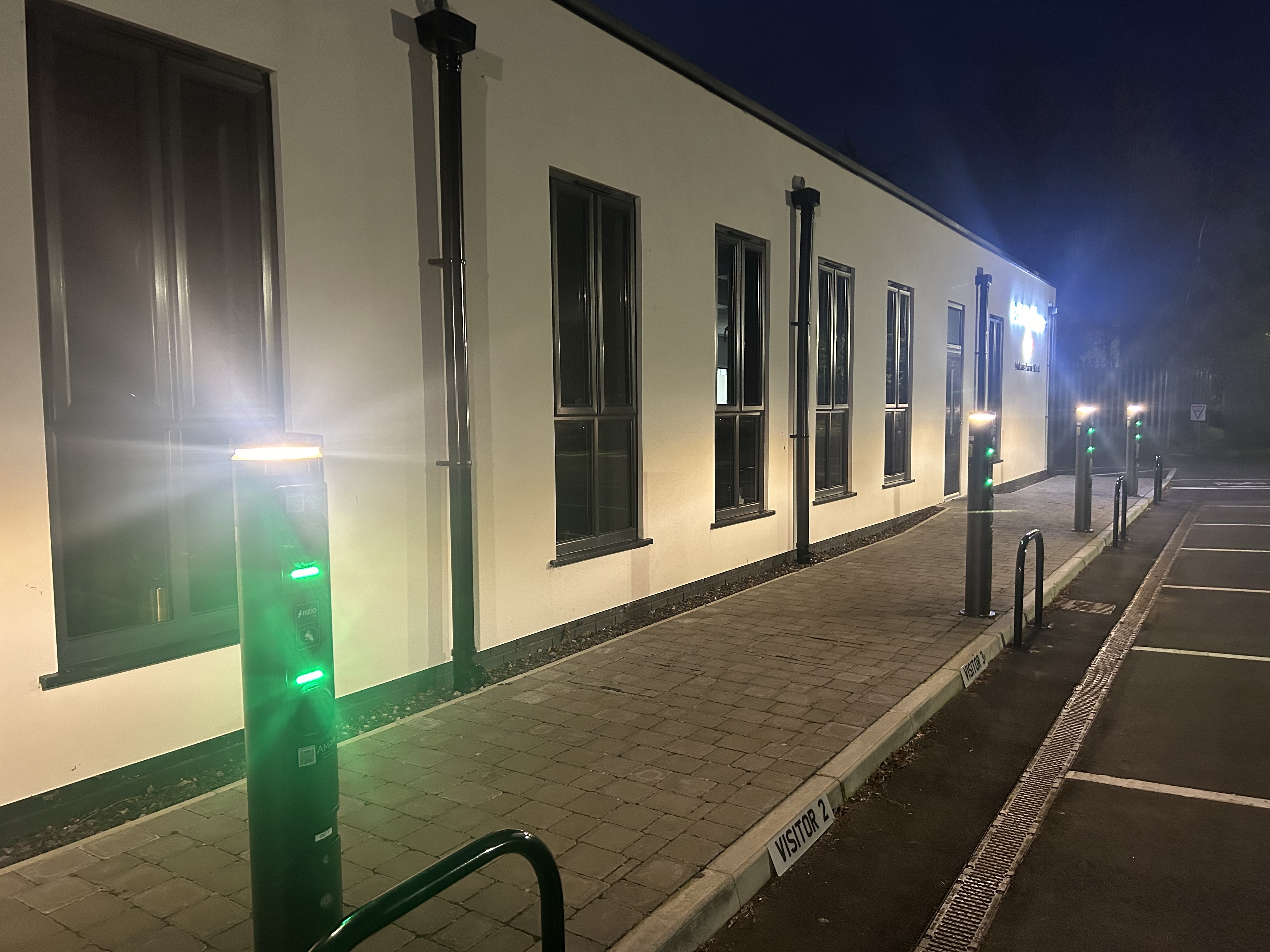 The Ratio io7 black charging pillar illuminated in ambient green light in front of a building.